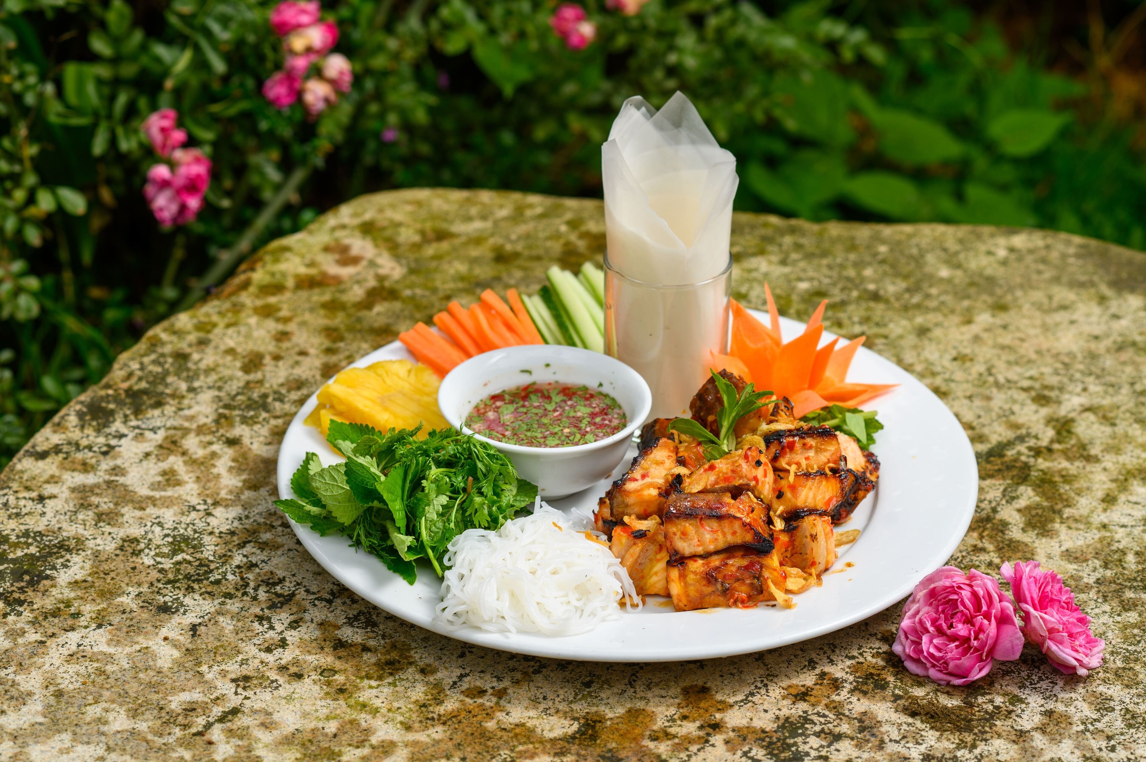 A plate of grilled chicken, vegetables, and noodles on a stone table with a drink and dipping sauce.