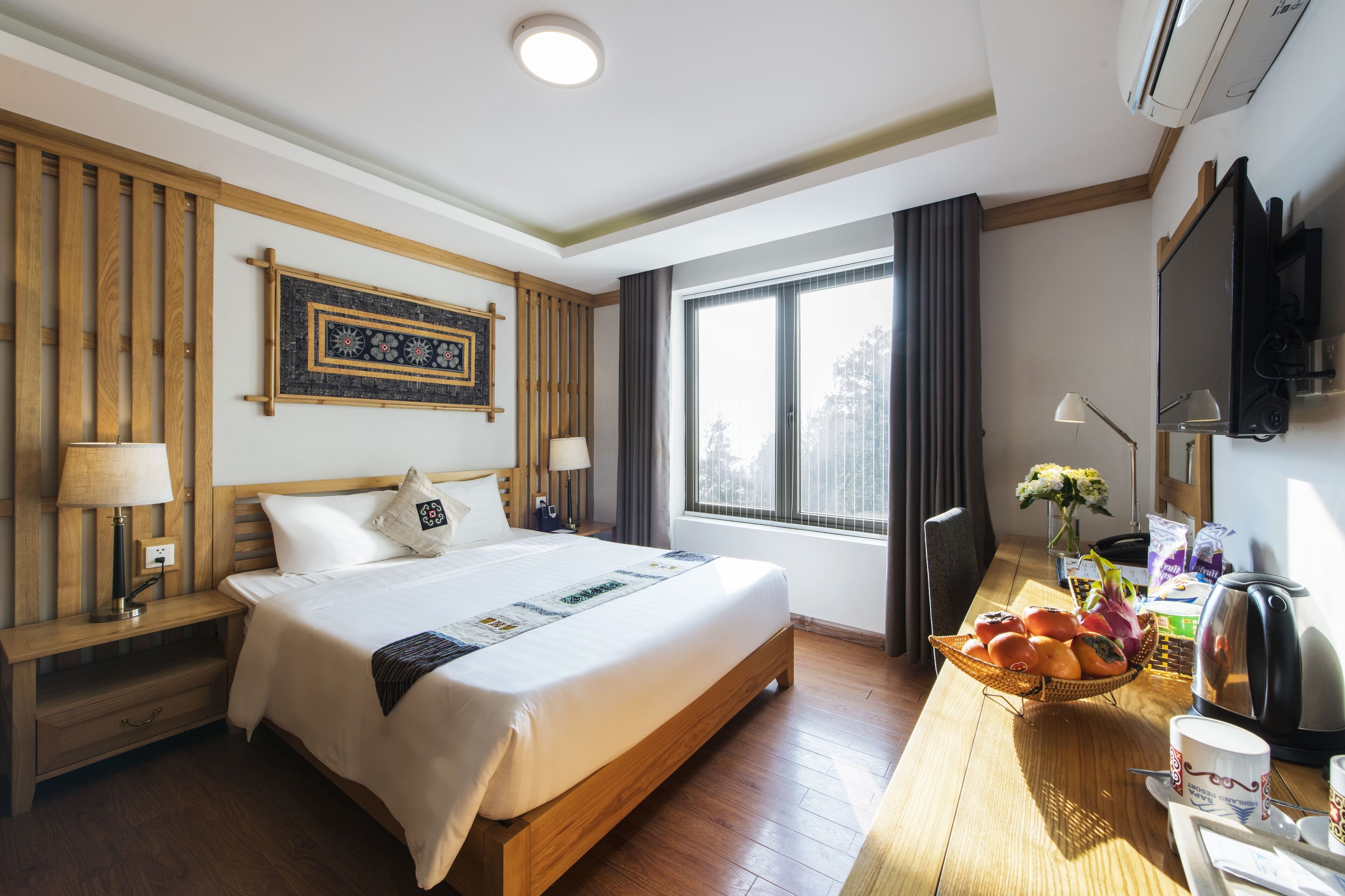 A hotel room with a large bed, wooden headboard, a flat-screen TV, a desk with a kettle, and a window with curtains.