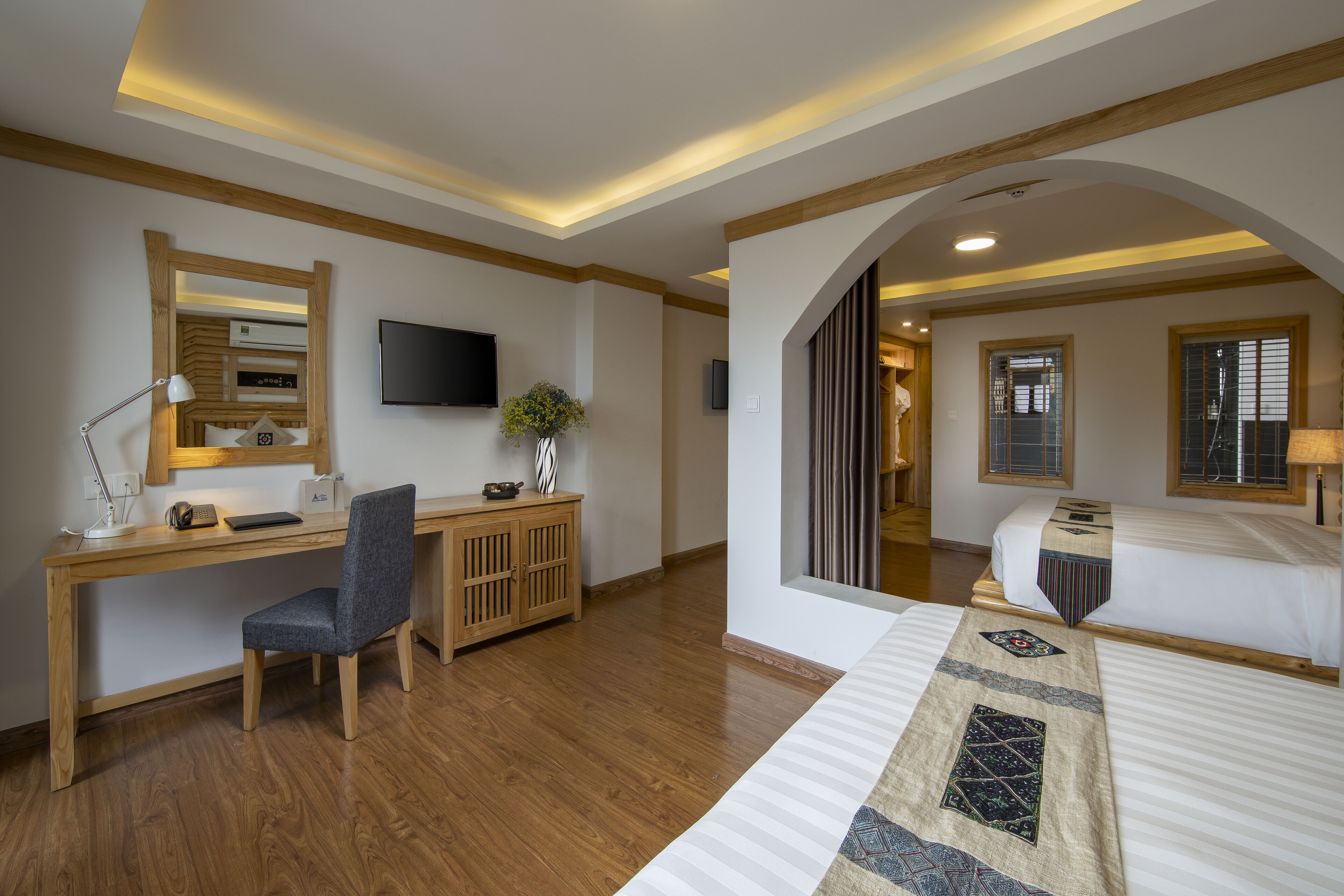 A hotel room with a wooden desk, a chair, a bed with a patterned bedspread, a television mounted on the wall, and a window with blinds.