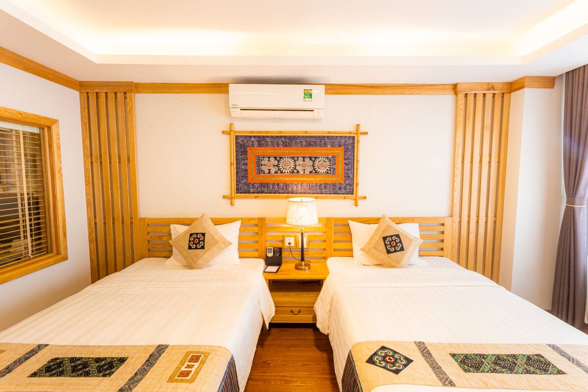 Ein Hotelzimmer mit zwei Betten, einem Holzkopfende, einer Wand-Klimaanlage, einem gemusterten Wandteppich und einem Nachttisch mit Lampe.