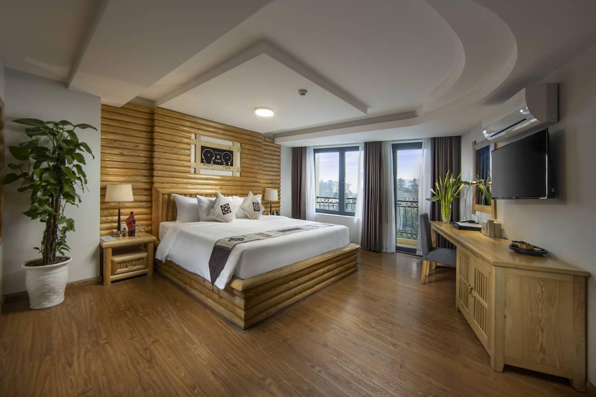 Ein geräumiges Hotelzimmer mit einem großen Bett, Holzvertäfelung, einem Fernseher, einem Balkon mit Vorhängen und einem Blumentopf.