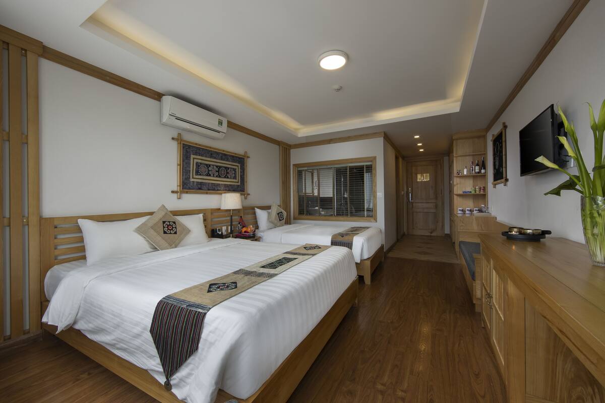Ein Hotelzimmer mit zwei Betten, Holzboden, Fernseher, einer Schrank mit Flaschen und einem Fenster mit Jalousien.