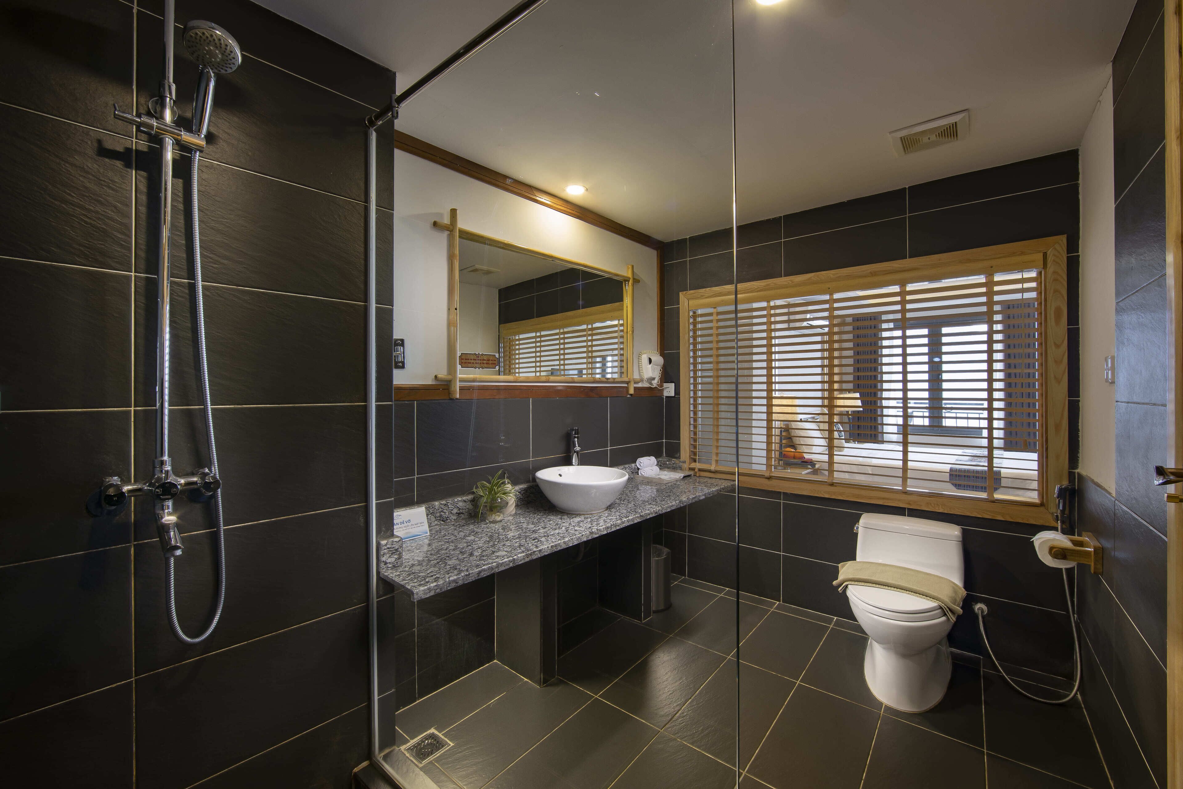 A modern bathroom with a glass-enclosed shower, a granite countertop sink, a toilet, and a window with wooden blinds.