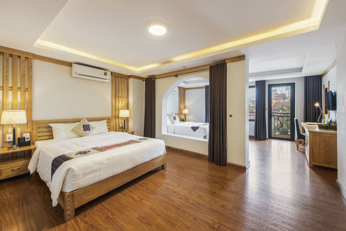Ein Hotelzimmer mit einem hölzernen Bett, Nachttischlampen, einer Klimaanlage, einem Balkon mit Blick und einem Schreibtisch mit Lampe.