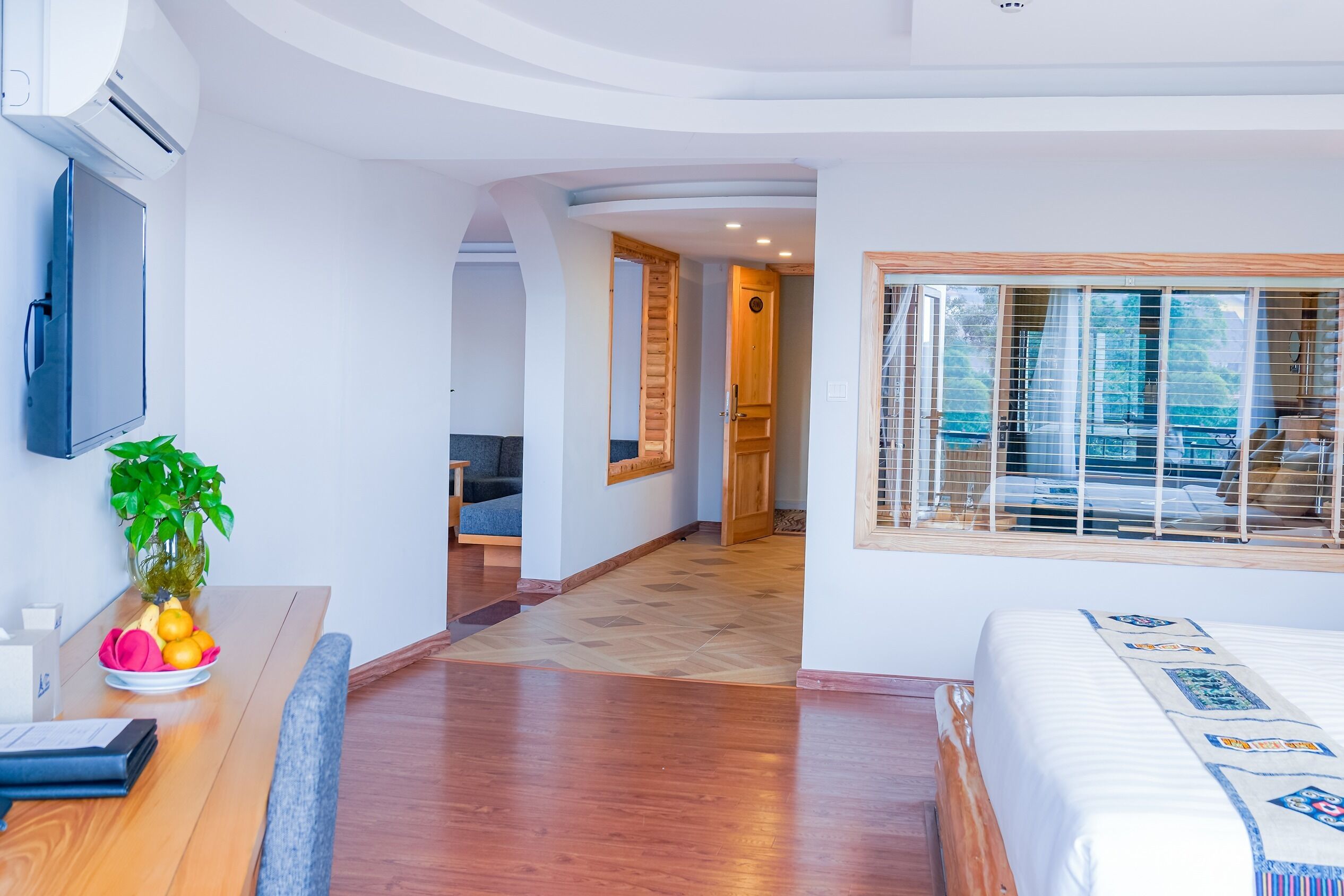 A modern hotel room with a wooden floor, a bed with a patterned bedspread, a TV mounted on the wall, and a window with blinds.