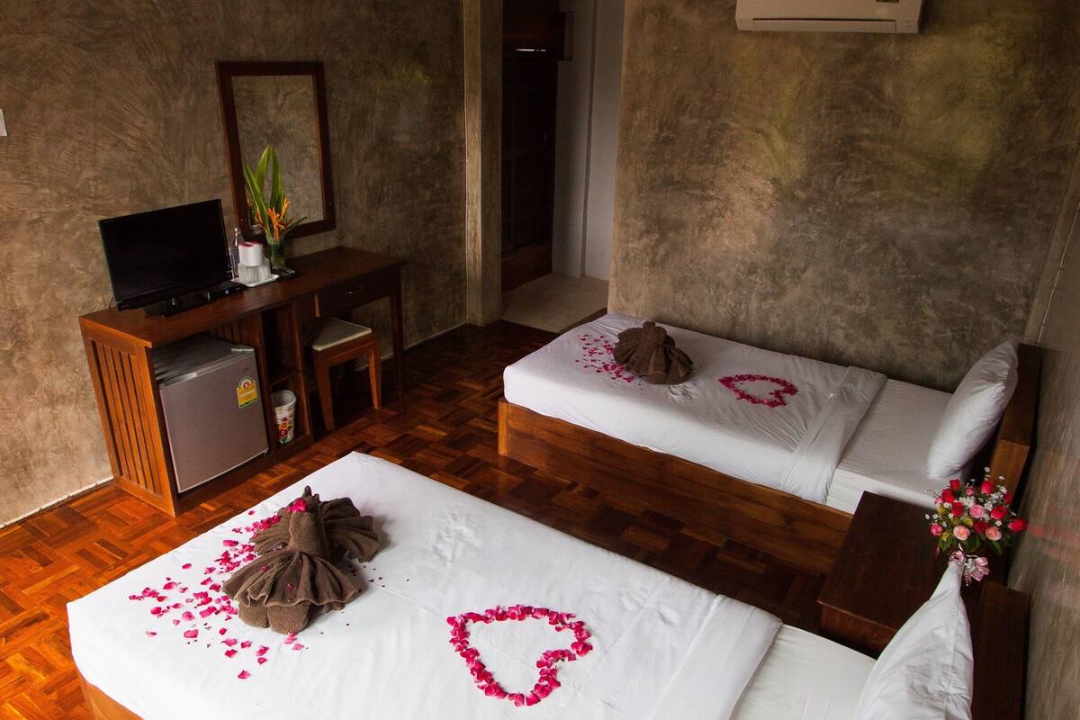 A room with two beds, a desk with a television, and an air conditioning unit.