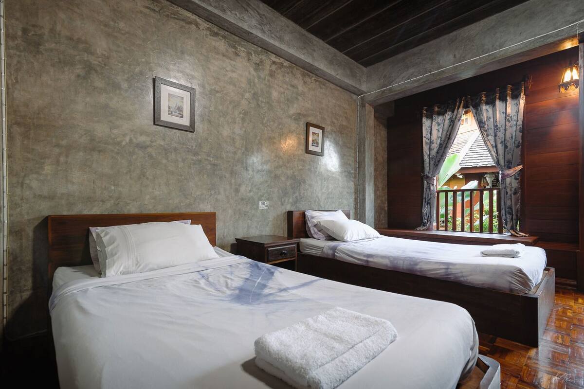 A room with two beds, a wooden headboard, a nightstand, a window with curtains, and a picture on the wall.