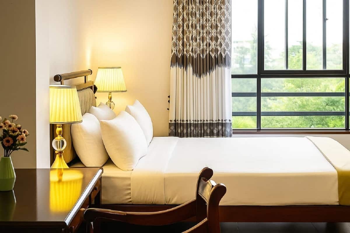 A hotel room with a bed, two bedside lamps, a chair, a vase with flowers, and a window with curtains.