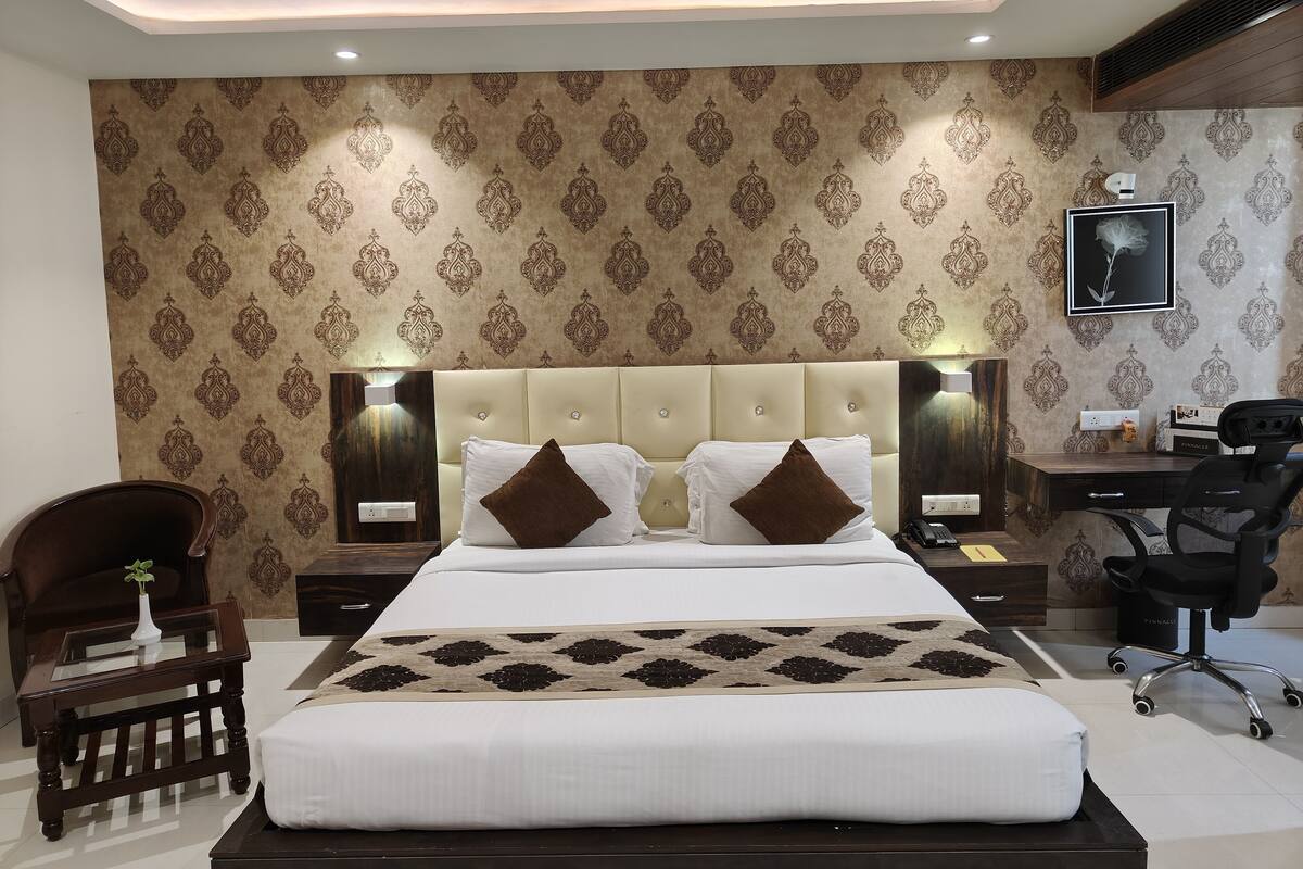 A modern hotel bedroom with a large bed, two bedside tables, a desk with a chair, and patterned wallpaper.