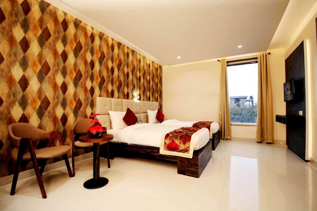 A hotel room with a bed, two chairs, a small table with flowers, a TV, and a window with curtains.