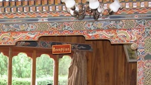 A reception desk with a sign labeled "Reception," a telephone, a vase with yellow flowers, and a window with a view of greenery. - Yangkhil Resort (Trongsa)