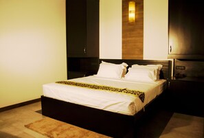 Een net opgemaakt bed met witte lakens en een geborduurde plaid, een nachtkastje met een lamp, en een donkere garderobe.