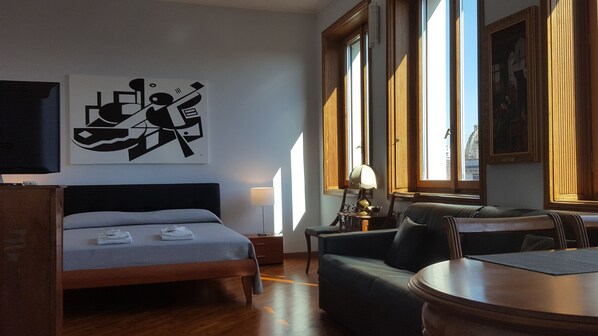 A hotel room with a bed, a desk, a chair, and a table. There is a painting on the wall and a window with a view of buildings.