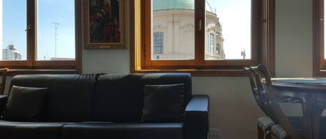 Une pièce comprenant un canapé en cuir noir, une chaise en bois et une table. De grandes fenêtres offrent une vue sur un bâtiment surmonté d’un dôme vert.