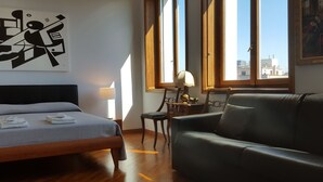 A hotel room with a bed, a leather sofa, a desk with a lamp, and a painting on the wall. - BnButler - Galleria Unione (Milan)