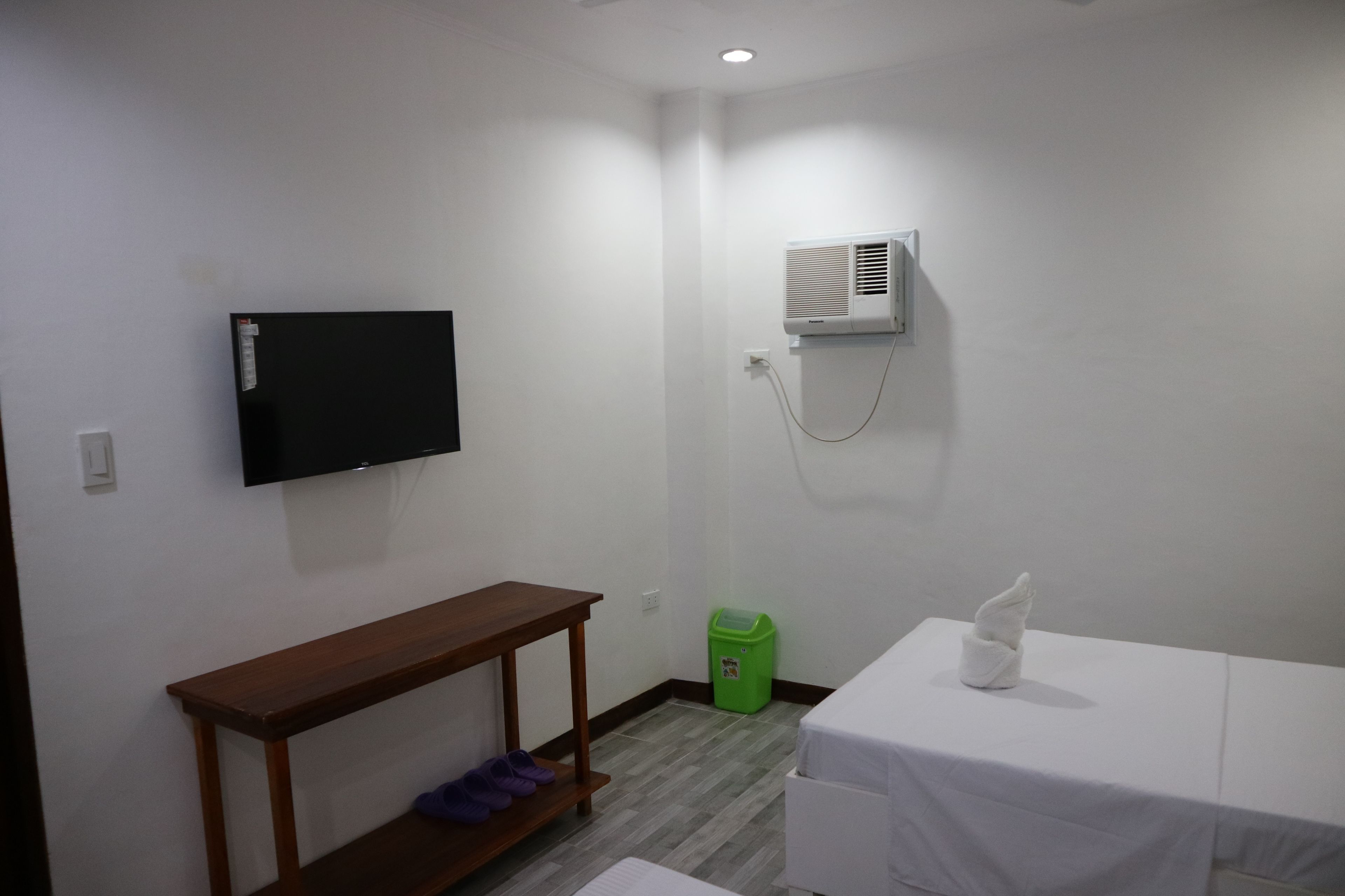 A small, clean room with a flat-screen TV mounted on the wall, a wooden bench, a small trash bin, and a table with a towel.