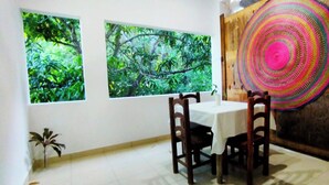 A dining area with a table and chairs, two large windows with a view of greenery, and a colorful woven wall hanging. - Yuu-lee Casa Bendita Huatulco (Santa María Huatulco)