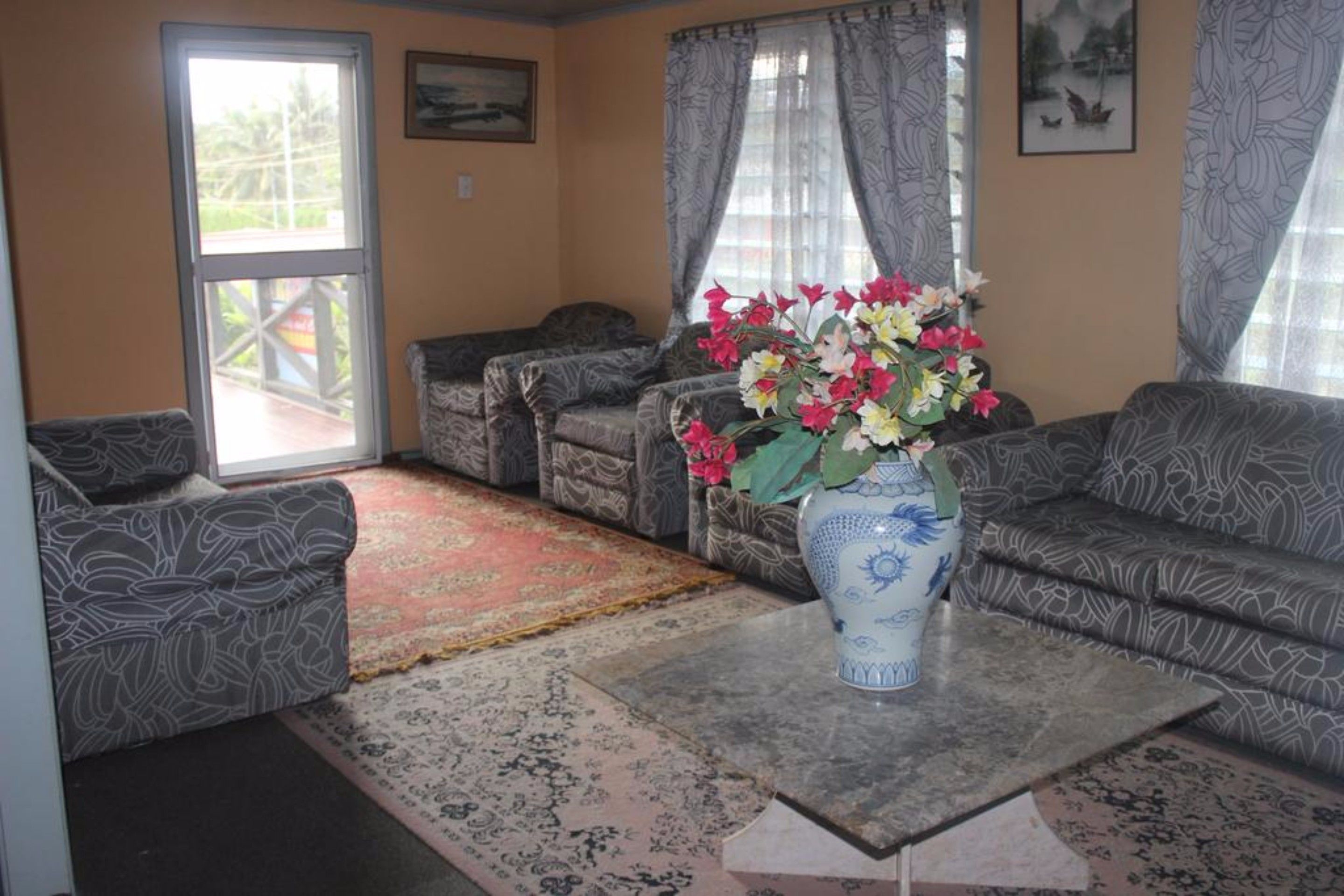 A living room with patterned sofas, a coffee table with a vase of flowers, and a large window with curtains.