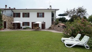 Uma casa de dois andares com um pátio que inclui uma mesa, cadeiras, um muro de pedra e um jardim com gramado e duas poltronas.