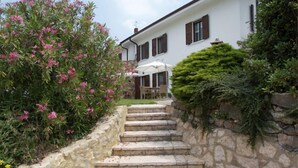 Uma escadaria de pedra leva a um edifício branco com persianas castanhas, cercado por vegetação e flores cor-de-rosa.