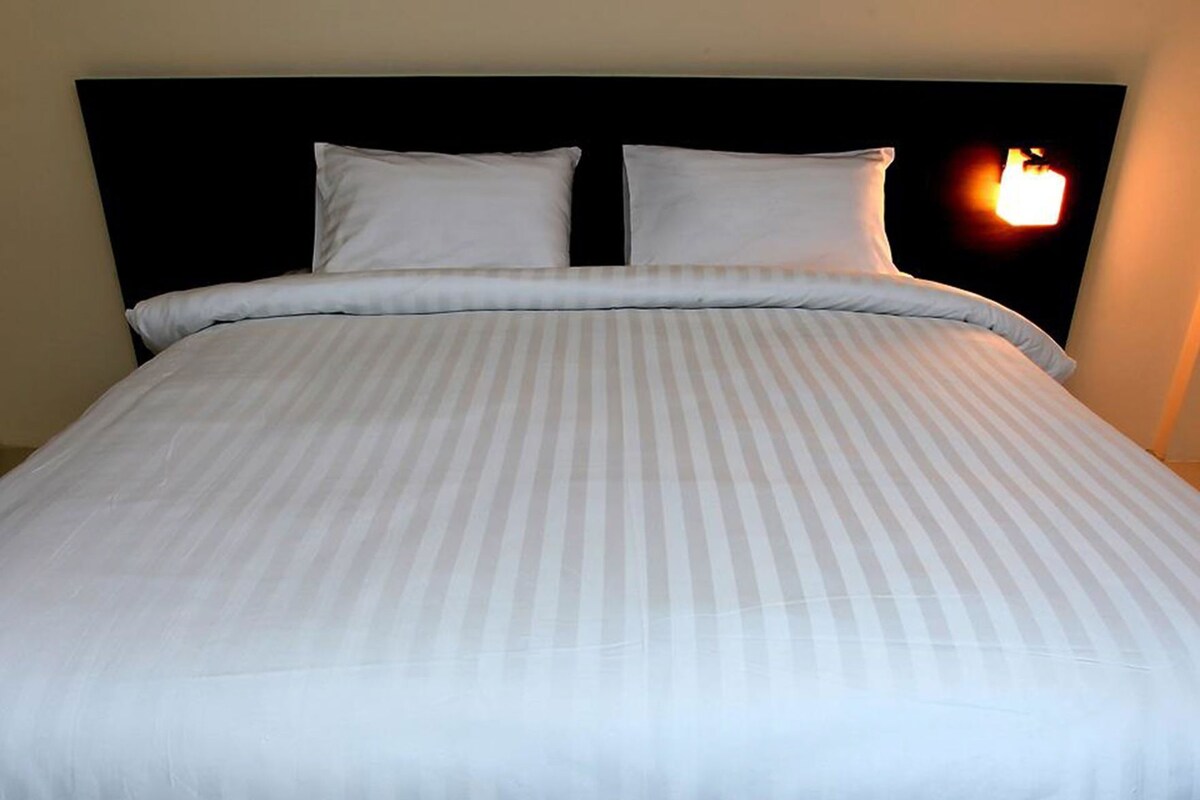Una cama con una colcha a rayas y dos almohadas, un cabecero negro, y una lámpara fijada a la pared.