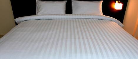 Una cama bien tendida con una colcha a rayas y dos almohadas, un cabecero negro y una lámpara fijada a la pared.