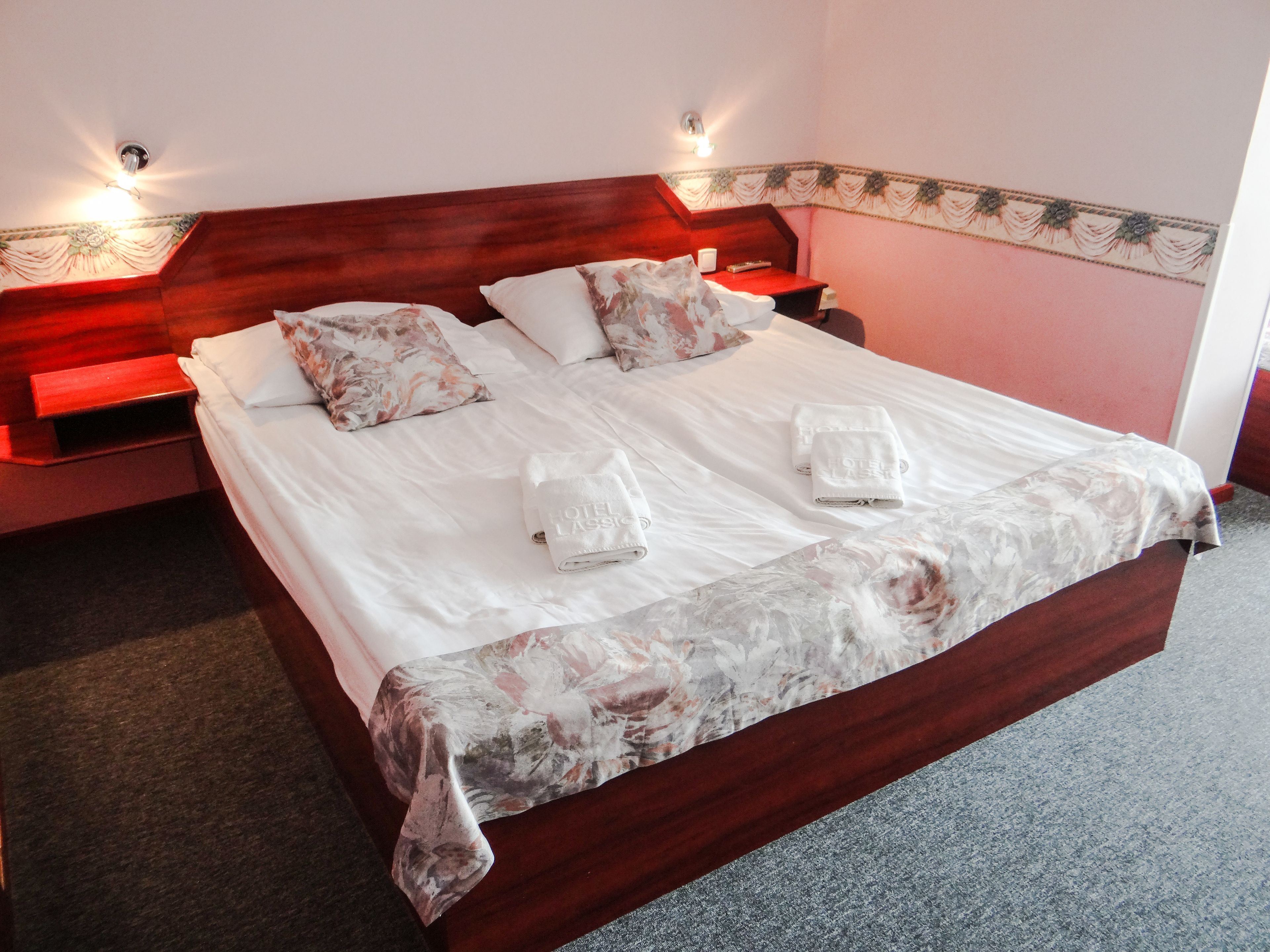 A neatly made bed with floral-patterned pillows and a white duvet, two folded towels on the bed, and a wooden headboard.
