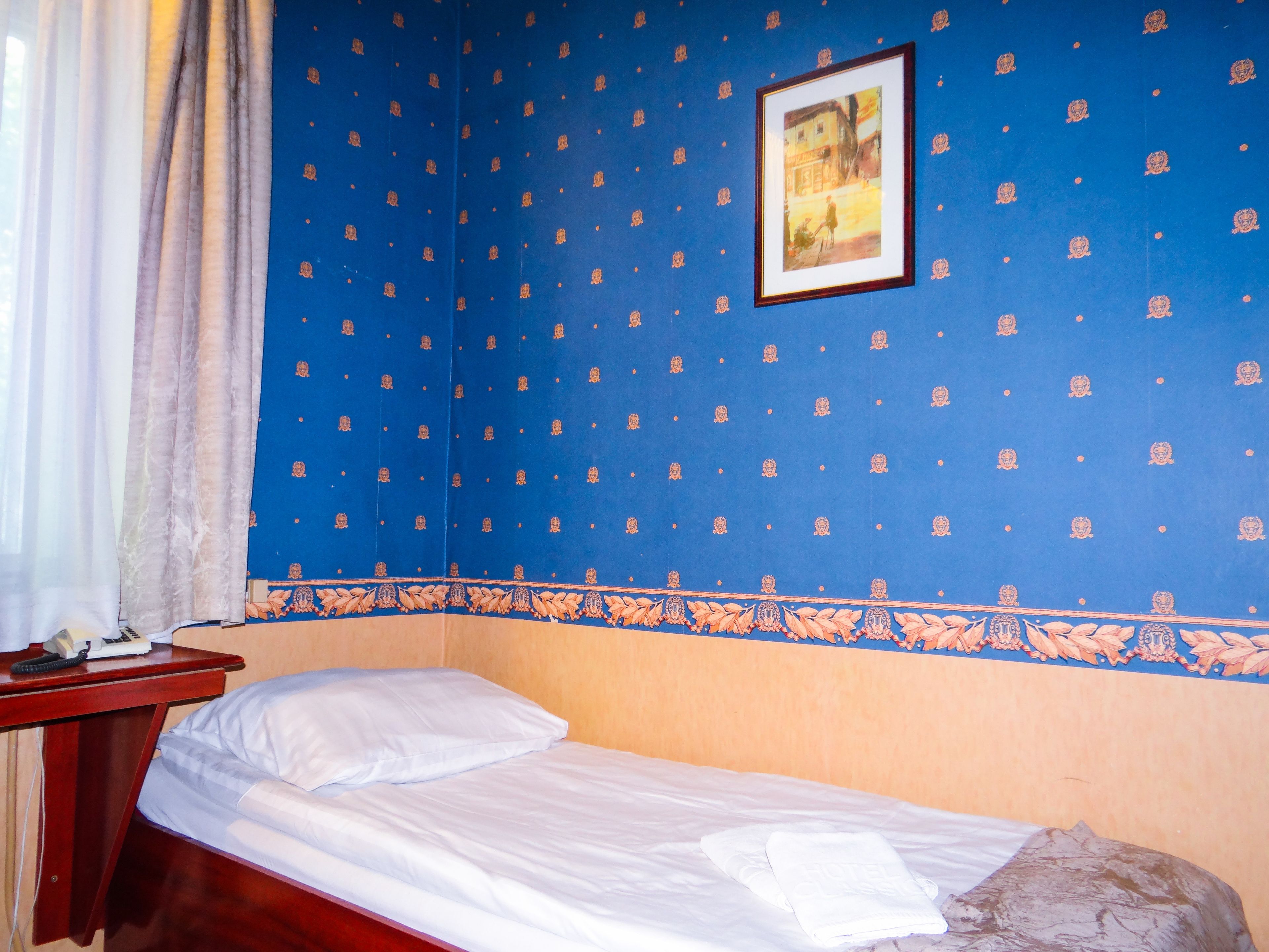 Una cama individual con ropa blanca, en una habitación con papel tapiz azul estampado y un cuadro en la pared.