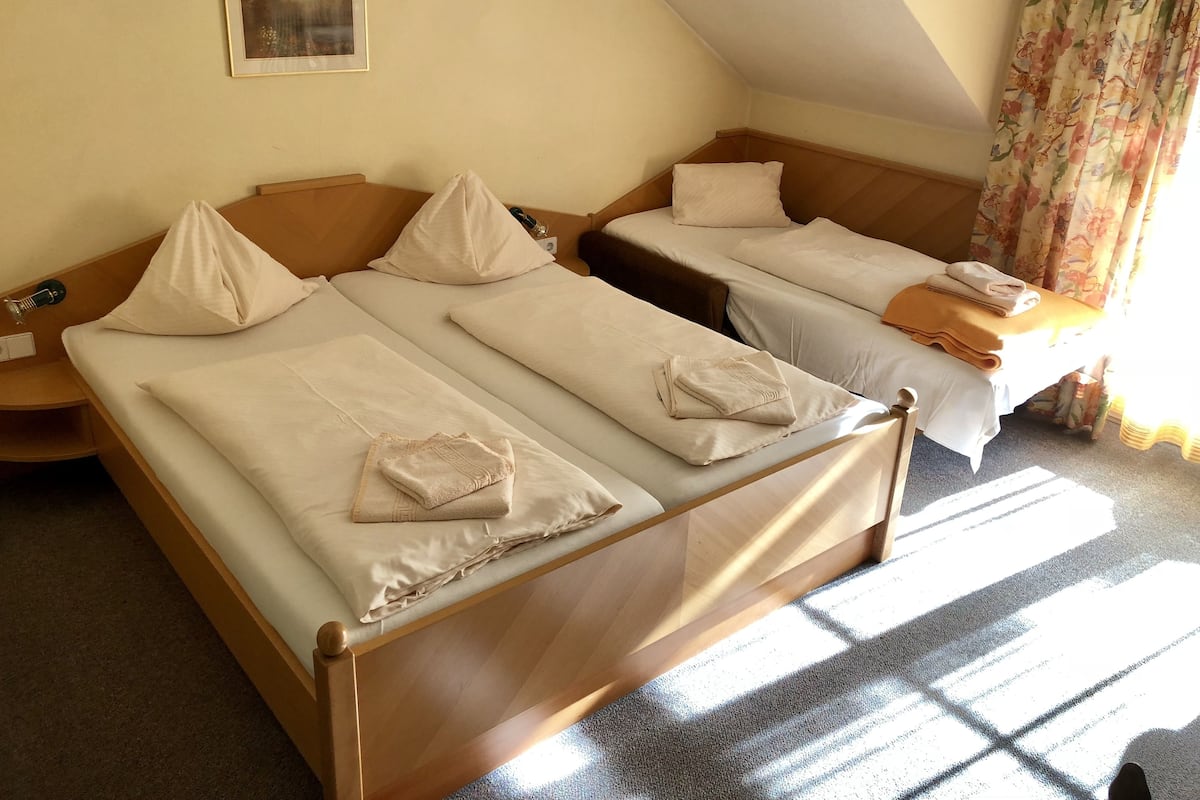 Une chambre d’hôtel avec deux lits simples, chacun muni d’un oreiller et d’une serviette pliée. On y trouve une table de chevet avec une lampe et une prise de courant. Un tableau encadré est accroché au mur, et une fenêtre avec des rideaux est visible.