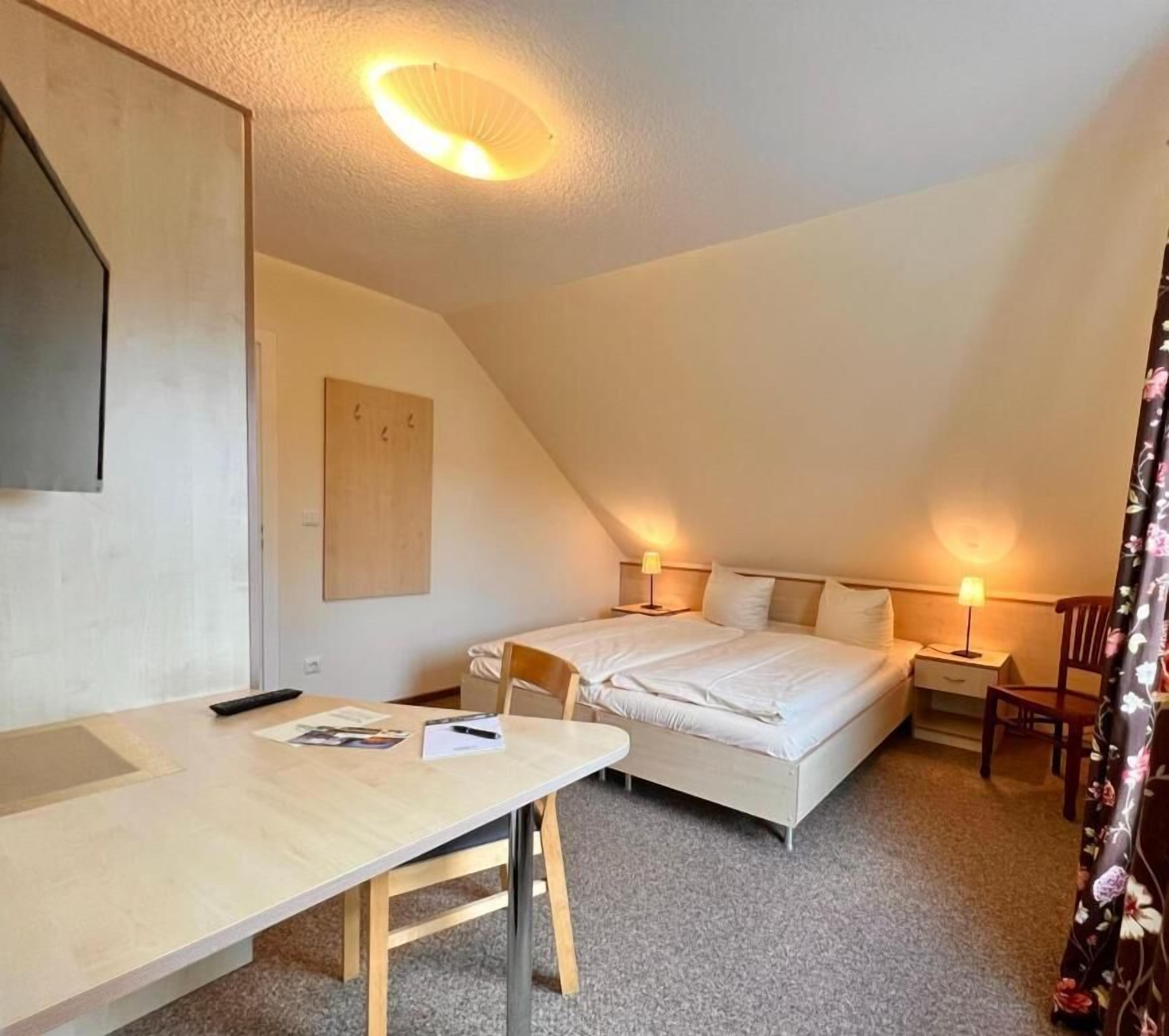 Habitació petita i acollidora amb un llit, una taula i una cadira. Hi ha dues làmpades de nit i una prestatgeria penjada a la paret.