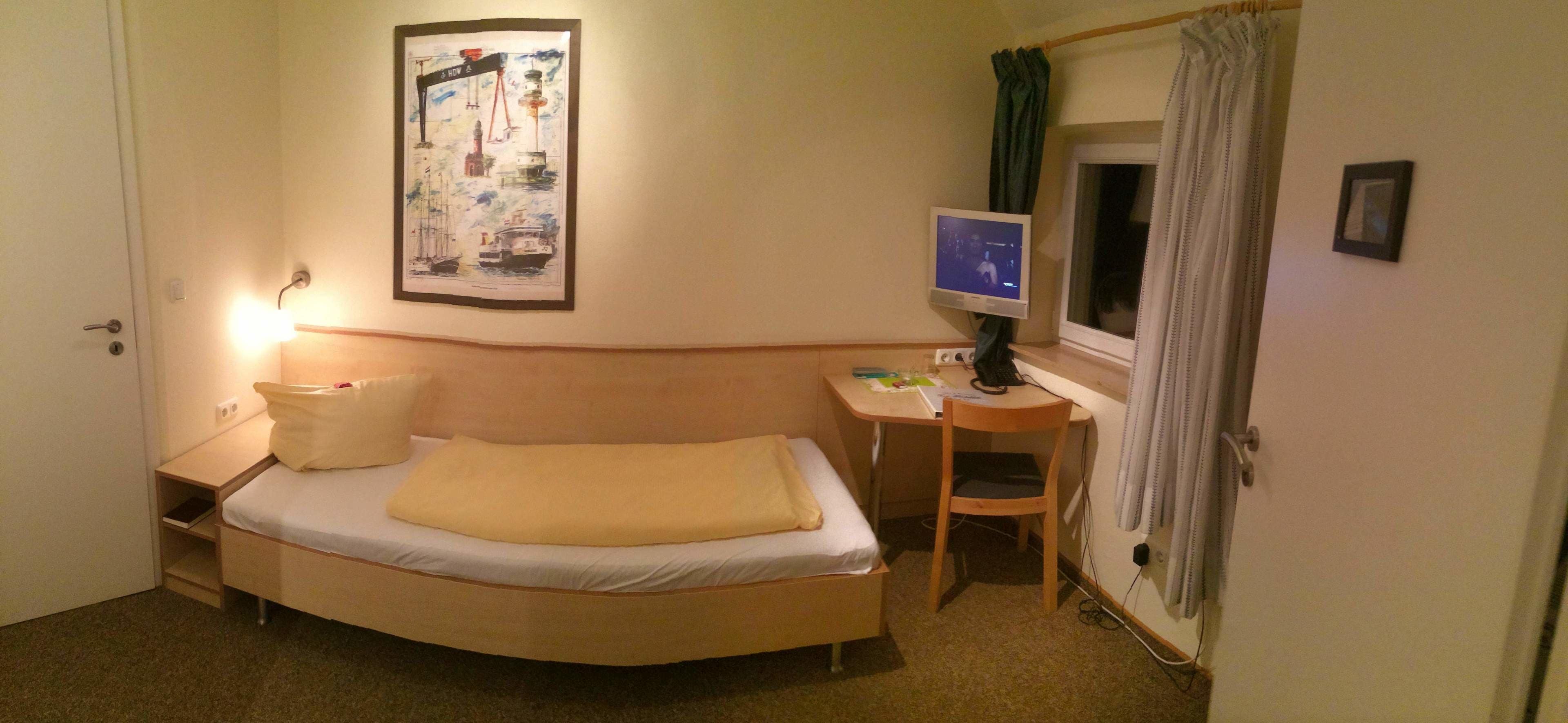 Ein kleines, ordentlich eingerichtetes Schlafzimmer mit Bett, Schreibtisch, Stuhl, Fernseher und einem Fenster mit Vorhängen.