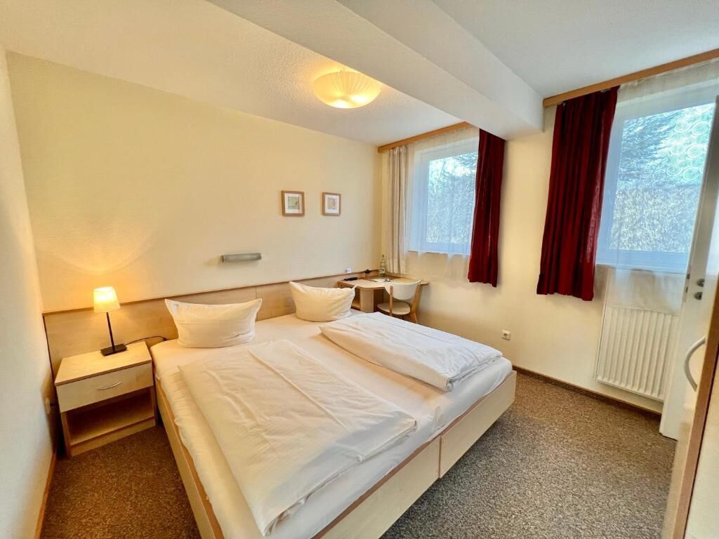 Una habitación de hotel con cama, mesita de noche, lámpara y una ventana con cortinas.