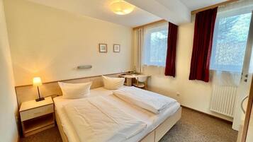 Una habitación de hotel con cama, mesita de noche, lámpara y una ventana con cortinas.