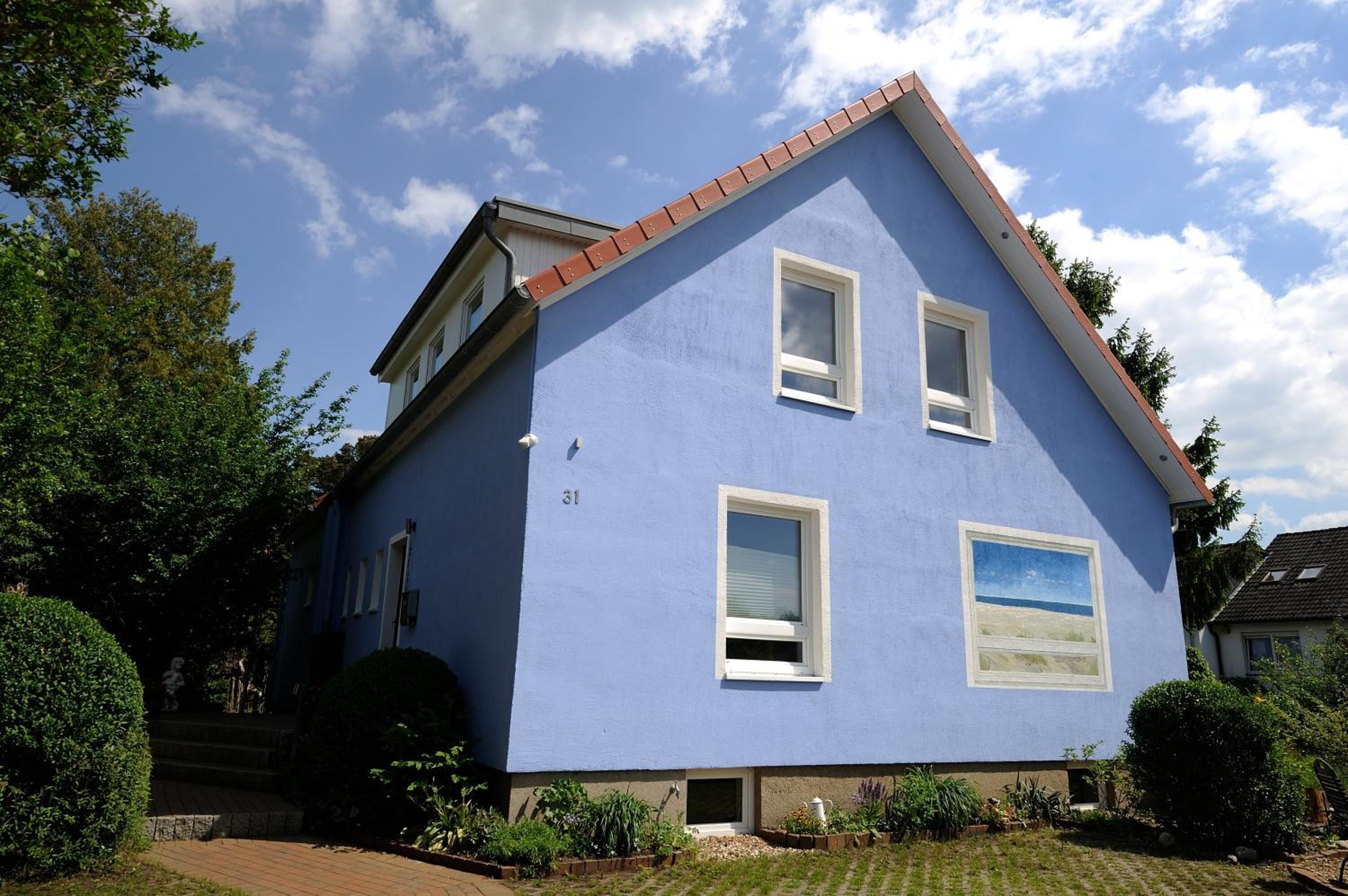 Uma casa de dois andares com fachada azul, número 31, com um pequeno jardim frontal e uma chaminé visível.