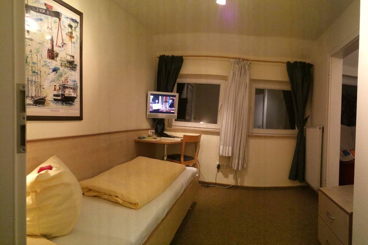 A small hotel room with a bed, a desk with a computer, a window with curtains, and a wall with a framed picture.