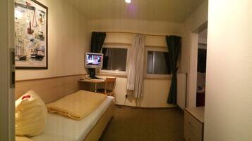 Una habitación de hotel pequeña con cama, un escritorio con computadora, una ventana con cortinas y una pared con un cuadro enmarcado.
