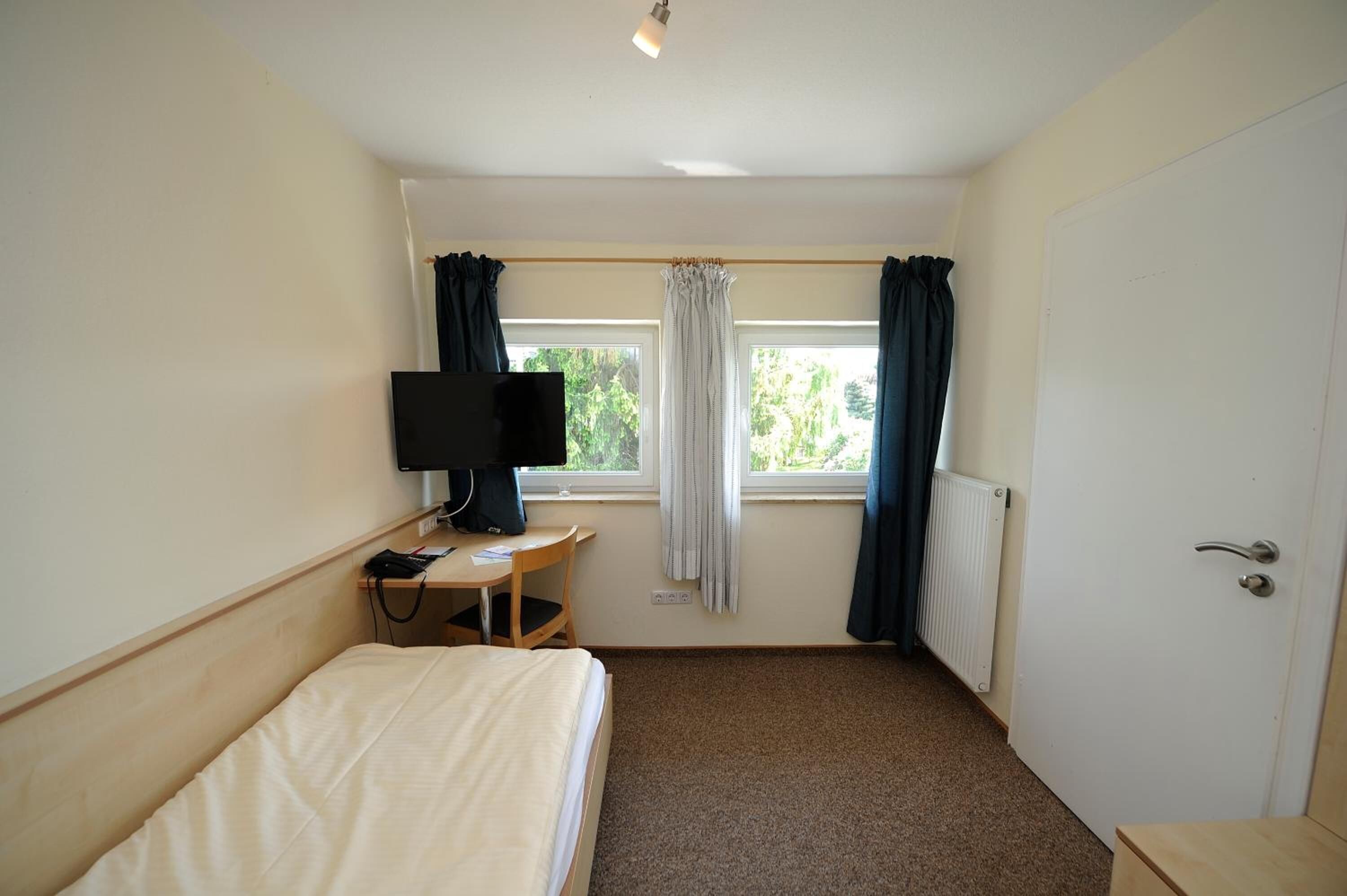 Quarto de hotel com cama, escrivaninha com televisão, janela com cortinas e radiador.