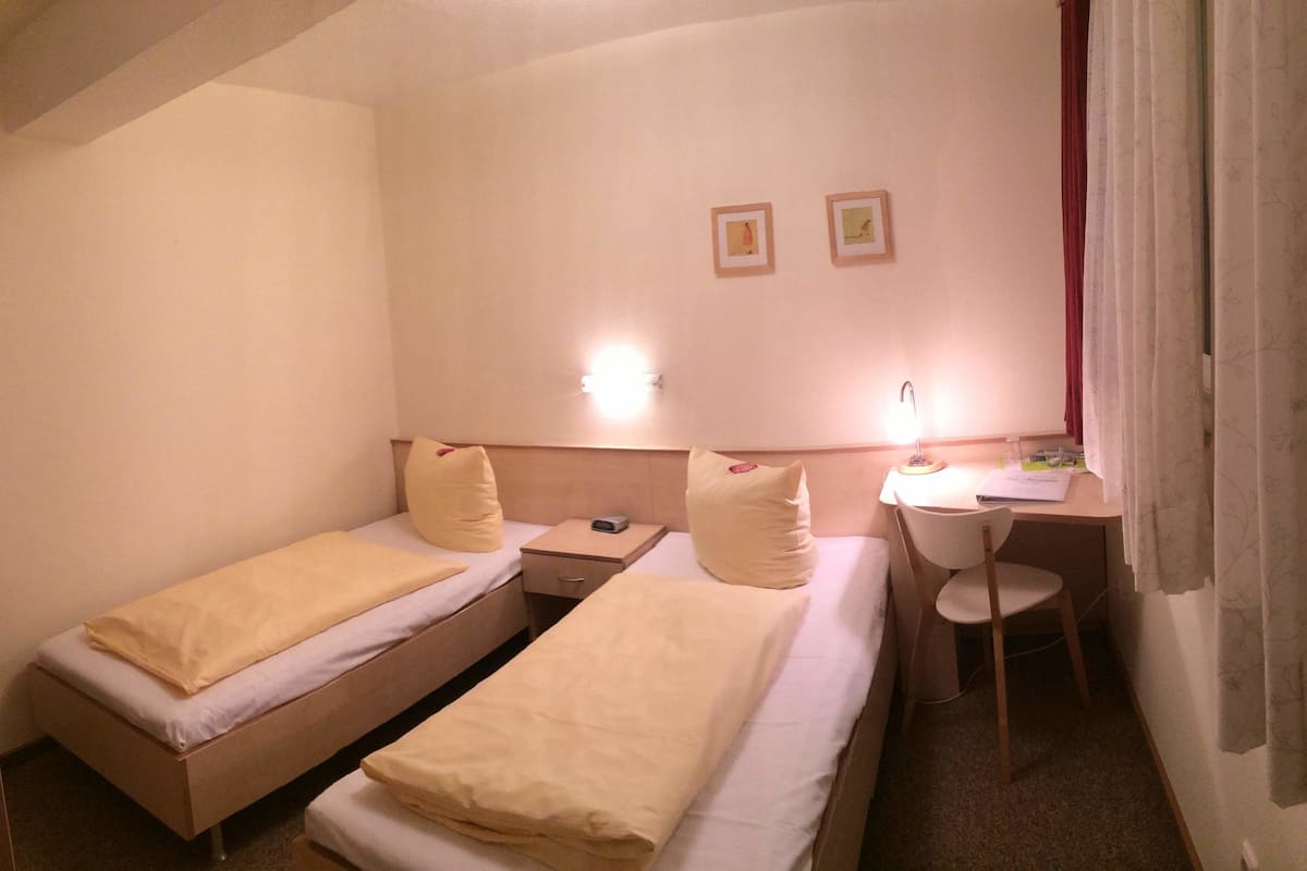 A small hotel room with two single beds, a desk, a chair, and a nightstand.