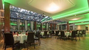 Un restaurante espacioso con numerosas mesas y sillas, ventanas amplias e iluminación ambiental verde.