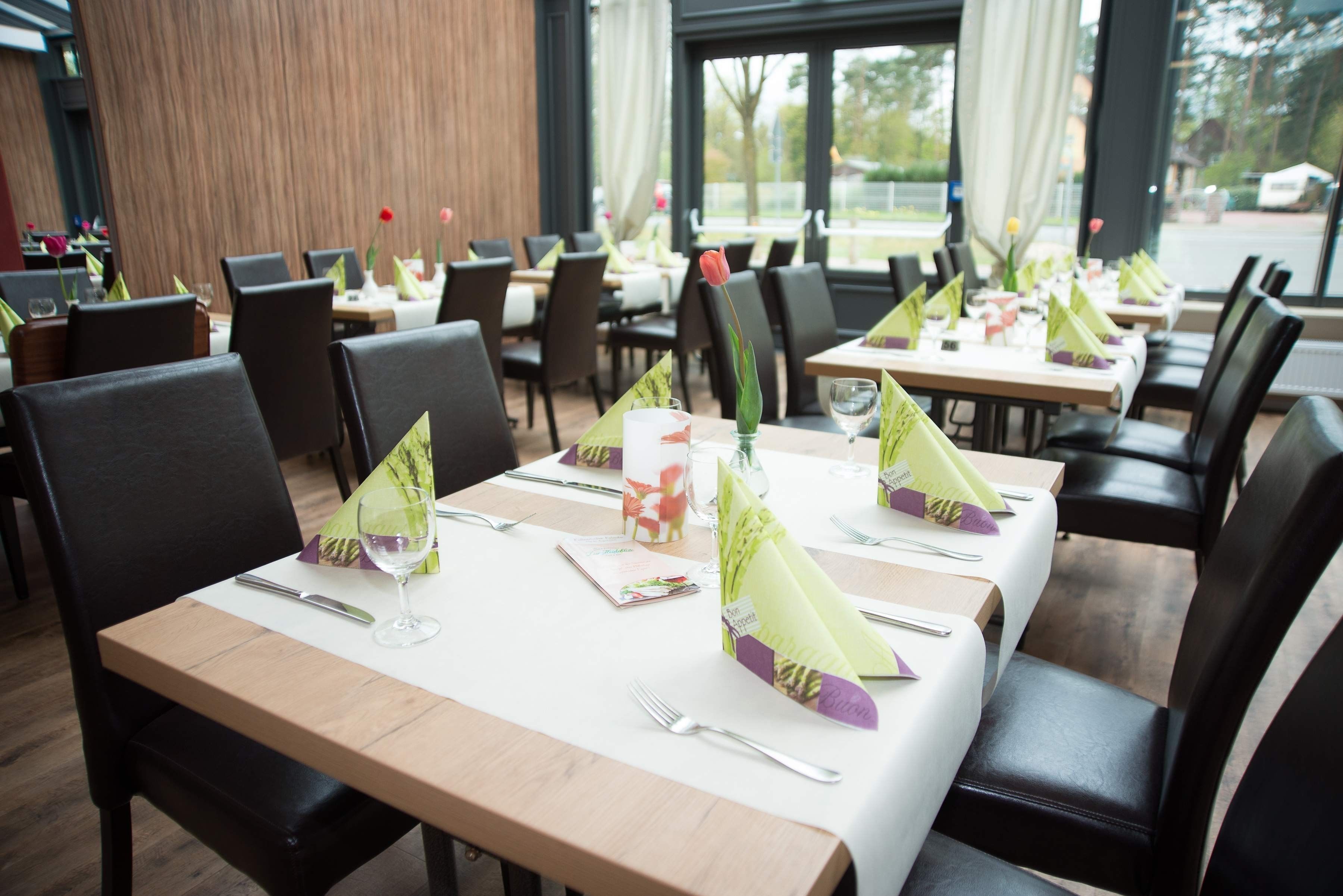 Un restaurant où les tables sont disposées avec soin, chacune étant recouverte d’une nappe blanche, agrémentée de serviettes vertes et de couverts en argent.