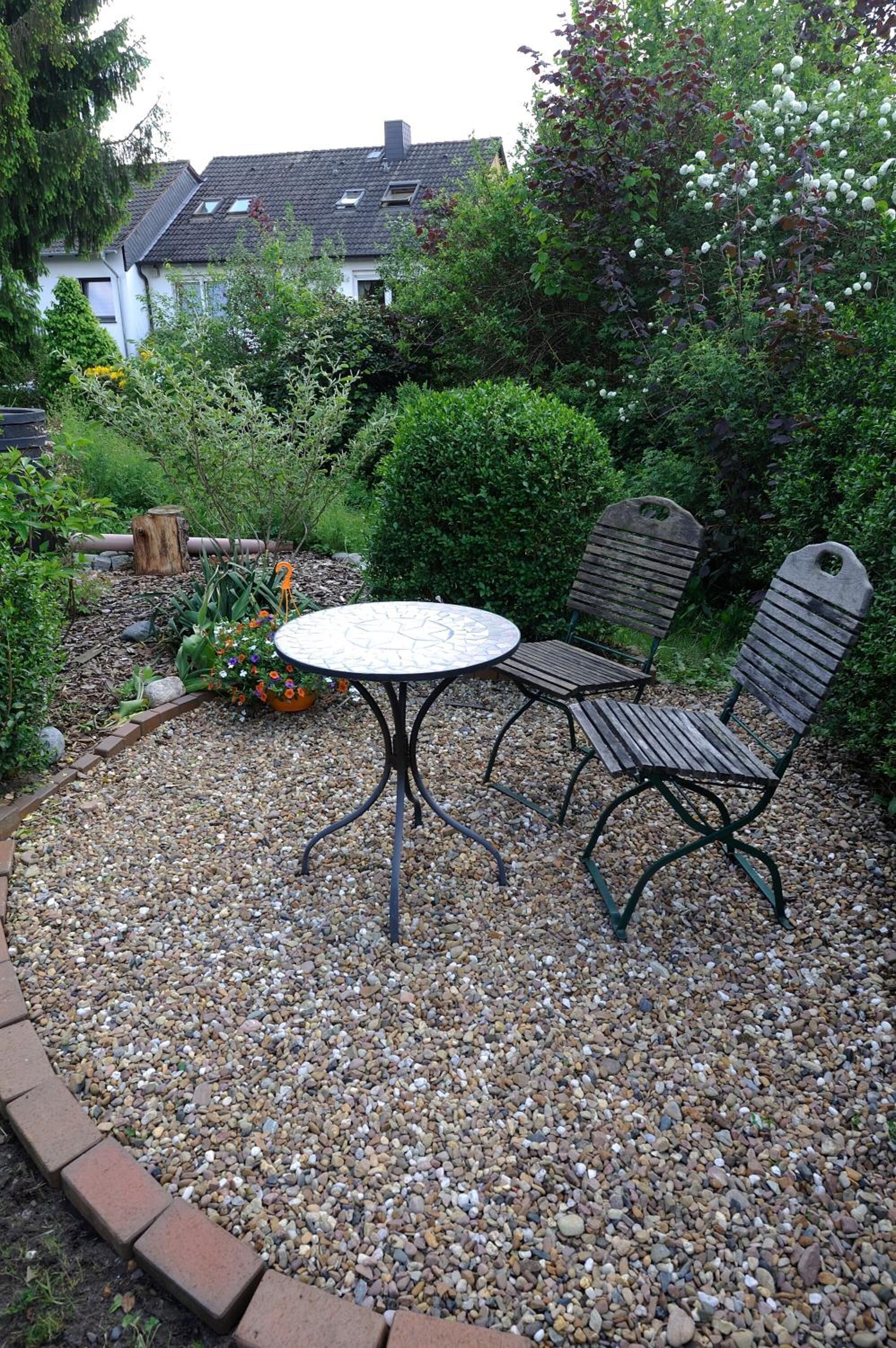 Une terrasse de jardin avec une table ronde et deux chaises en bois, entourée de gravier et de plantes.