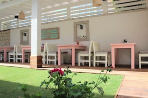 An outdoor patio area with white and pink furniture, including tables and chairs, under a white pergola. There are potted plants and a small garden area in the foreground. - Villamer Hotel Alacati (Cesme)