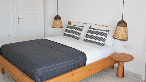 Una cama con un colchón gris oscuro, ropa de cama blanca y almohadas a rayas. Junto a la cama hay un taburete de madera.