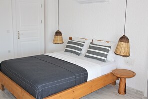 A bed with a dark grey mattress, white bedding, and striped pillows. There is a small wooden stool beside the bed.