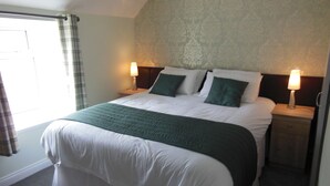 Een net opgemaakt bed met een groen-wit sprei, twee bedlampjes en een raam met gordijnen.