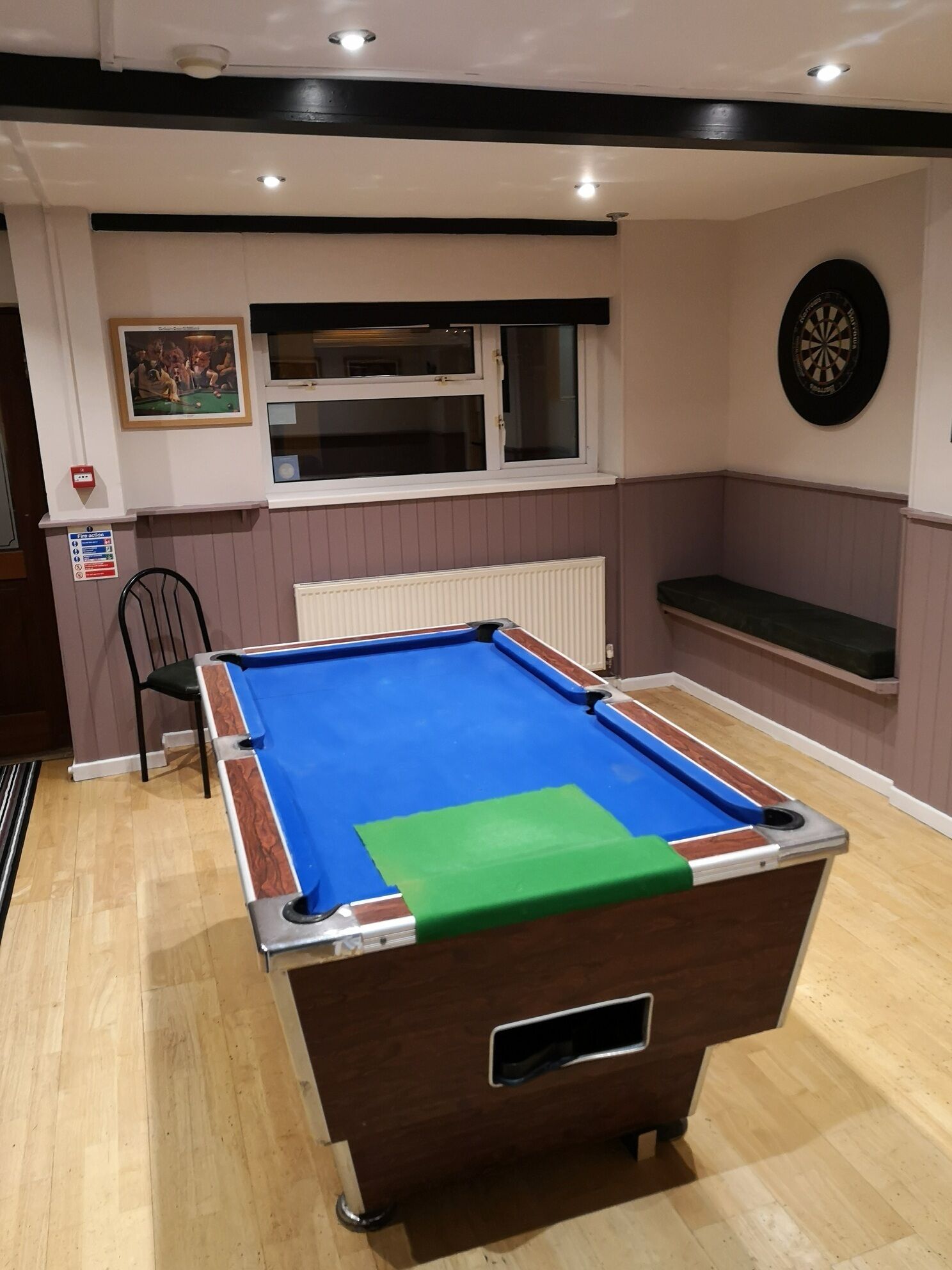 Ein Billardtisch mit blauer Spielfläche und grüner Tischdecke, ein Dartboard, ein gerahmtes Bild und ein Fenster mit Jalousien.