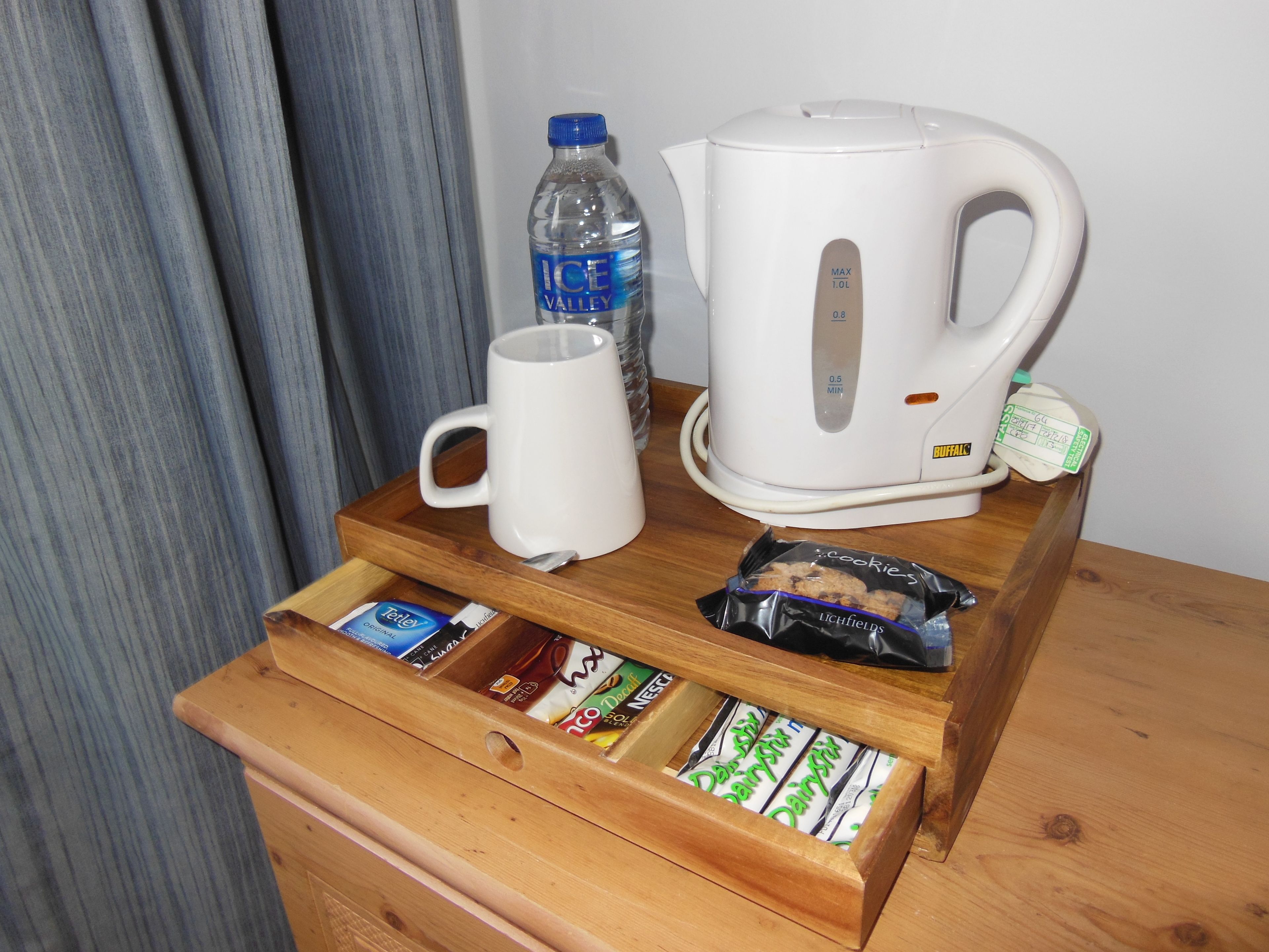 Ein Holztablett mit einer weißen Tasse, einer Wasserflasche, einer Kaffeemaschine und einer Schublade mit Tee- und Keksbeuteln.