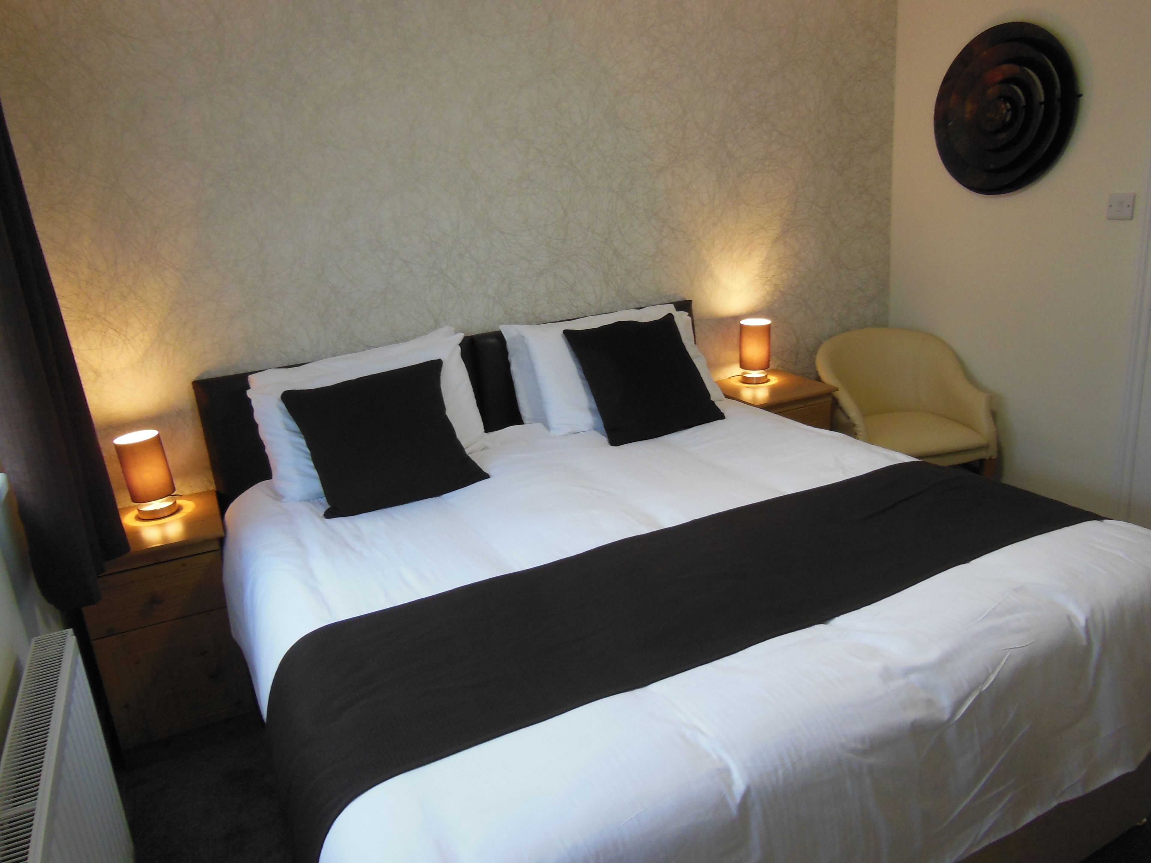 A neatly made bed with black and white bedding, two bedside lamps, and a chair in a room with a plain wall and a radiator.