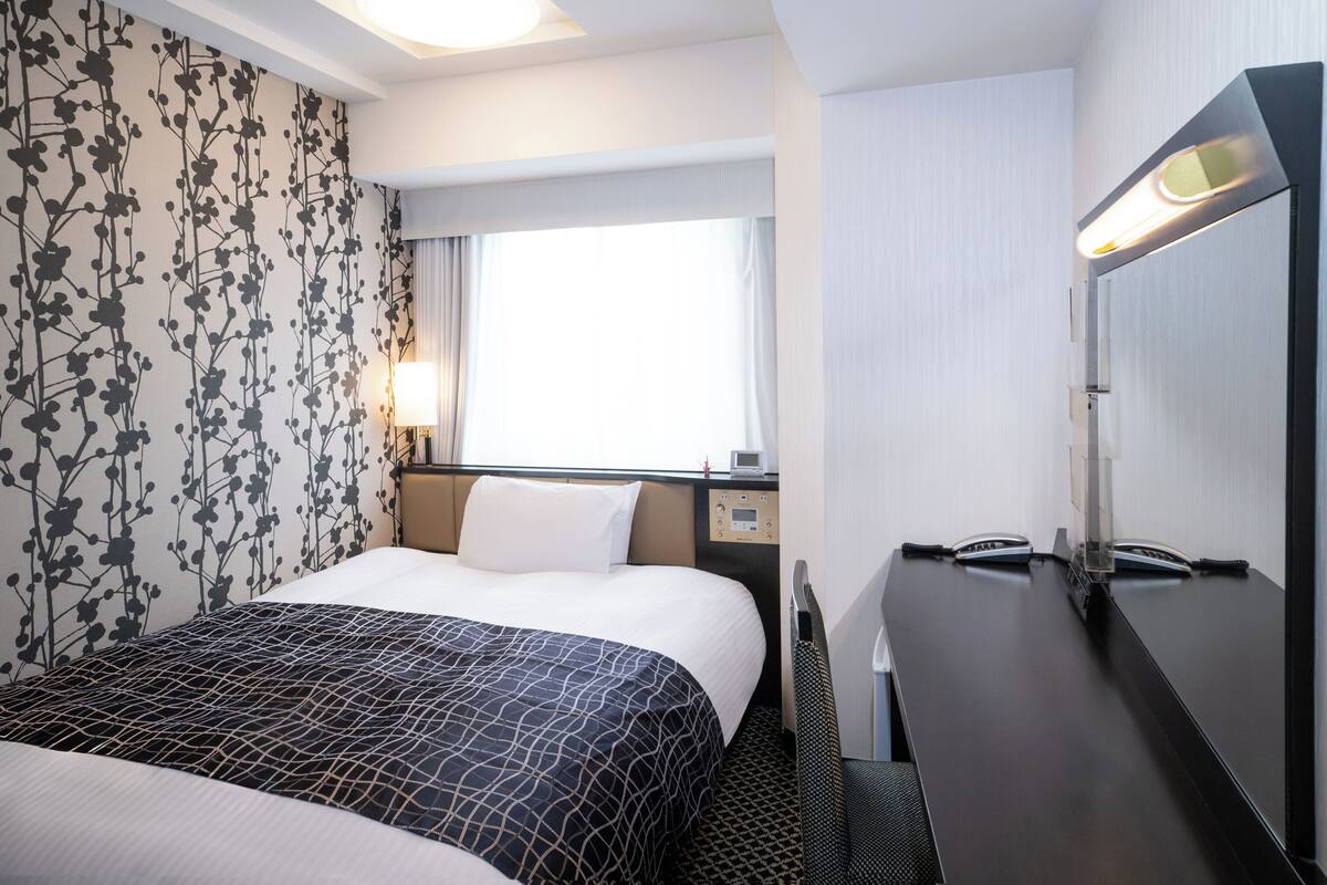 A hotel room with a bed, a desk, a chair, a lamp, and a window.