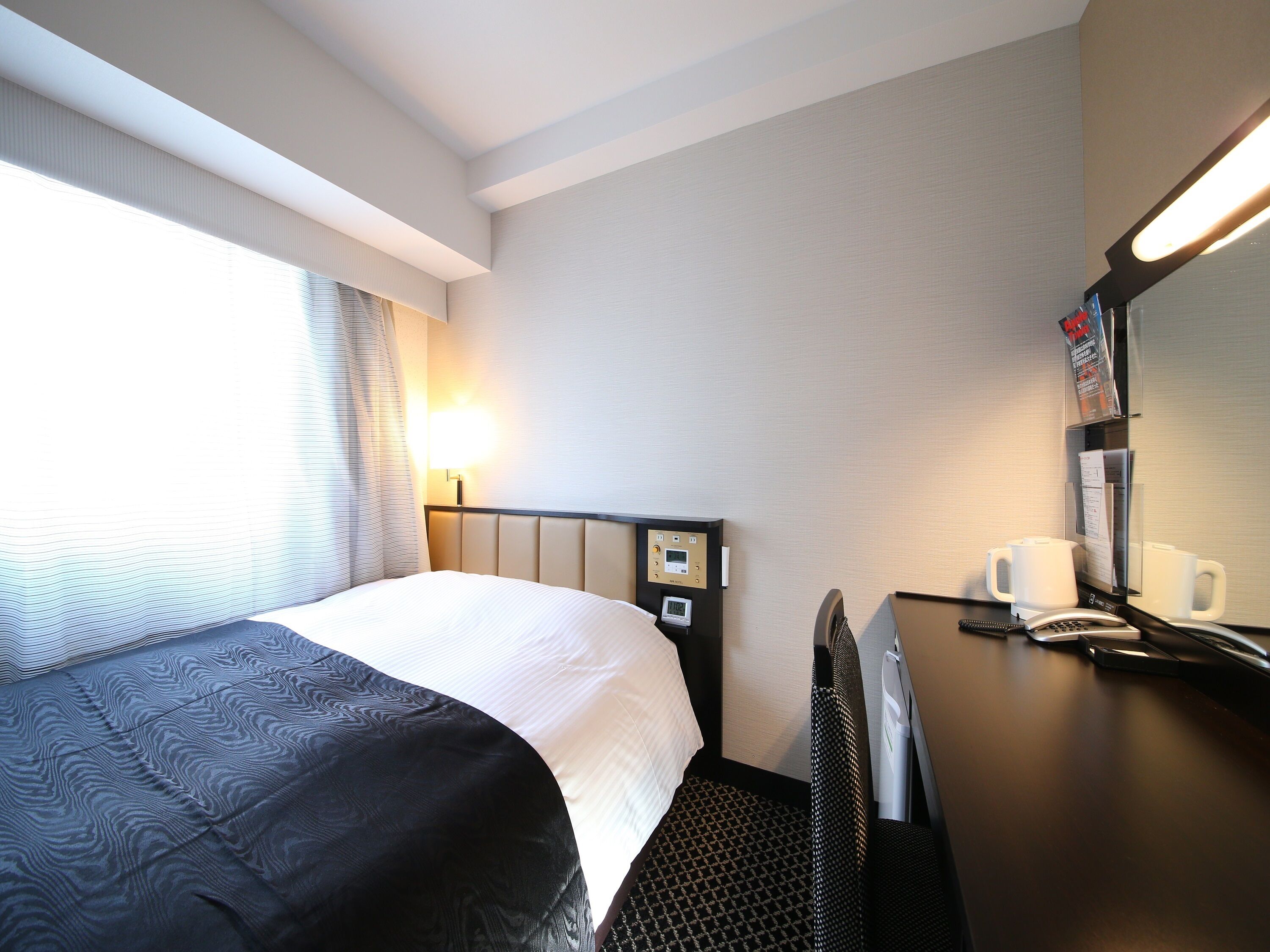 A hotel room with a bed, a desk with a telephone and a coffee maker, a headboard with controls, and a window with curtains.